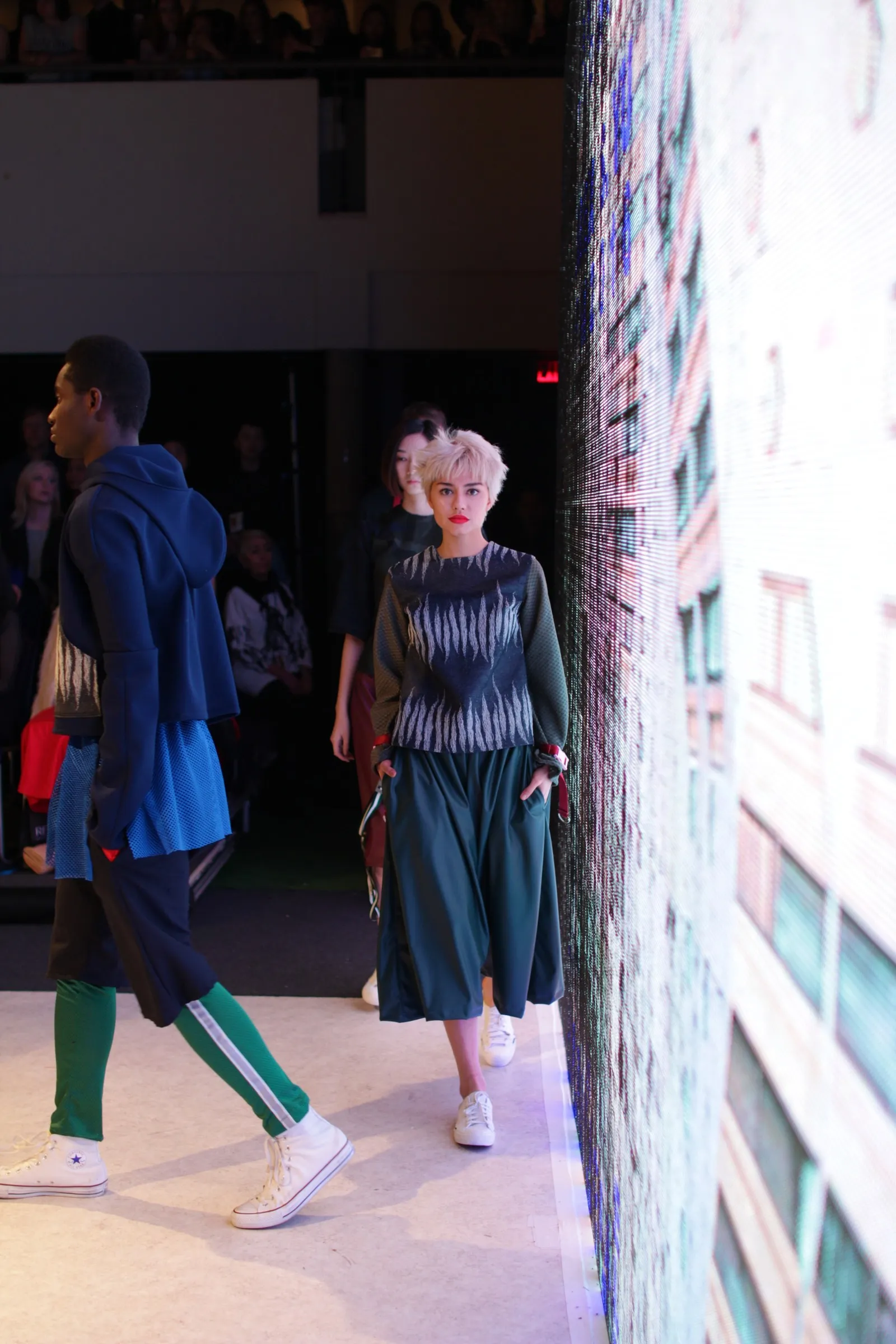 Models walking a runway past a wall of screens at a fashion show