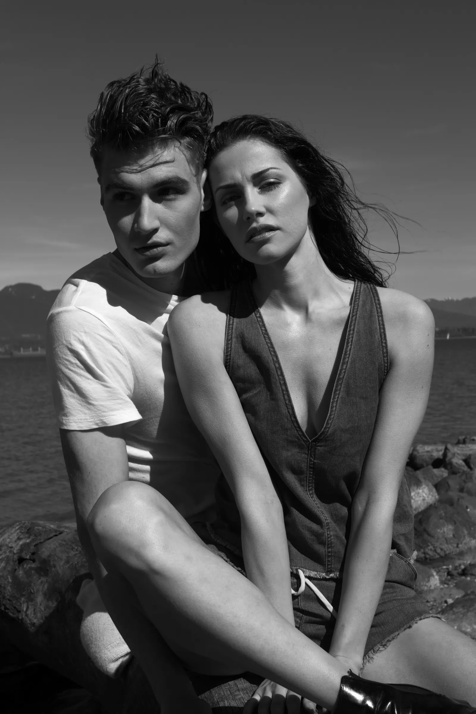 Black-and-white portrait of a young couple sitting close together on rocks by the water