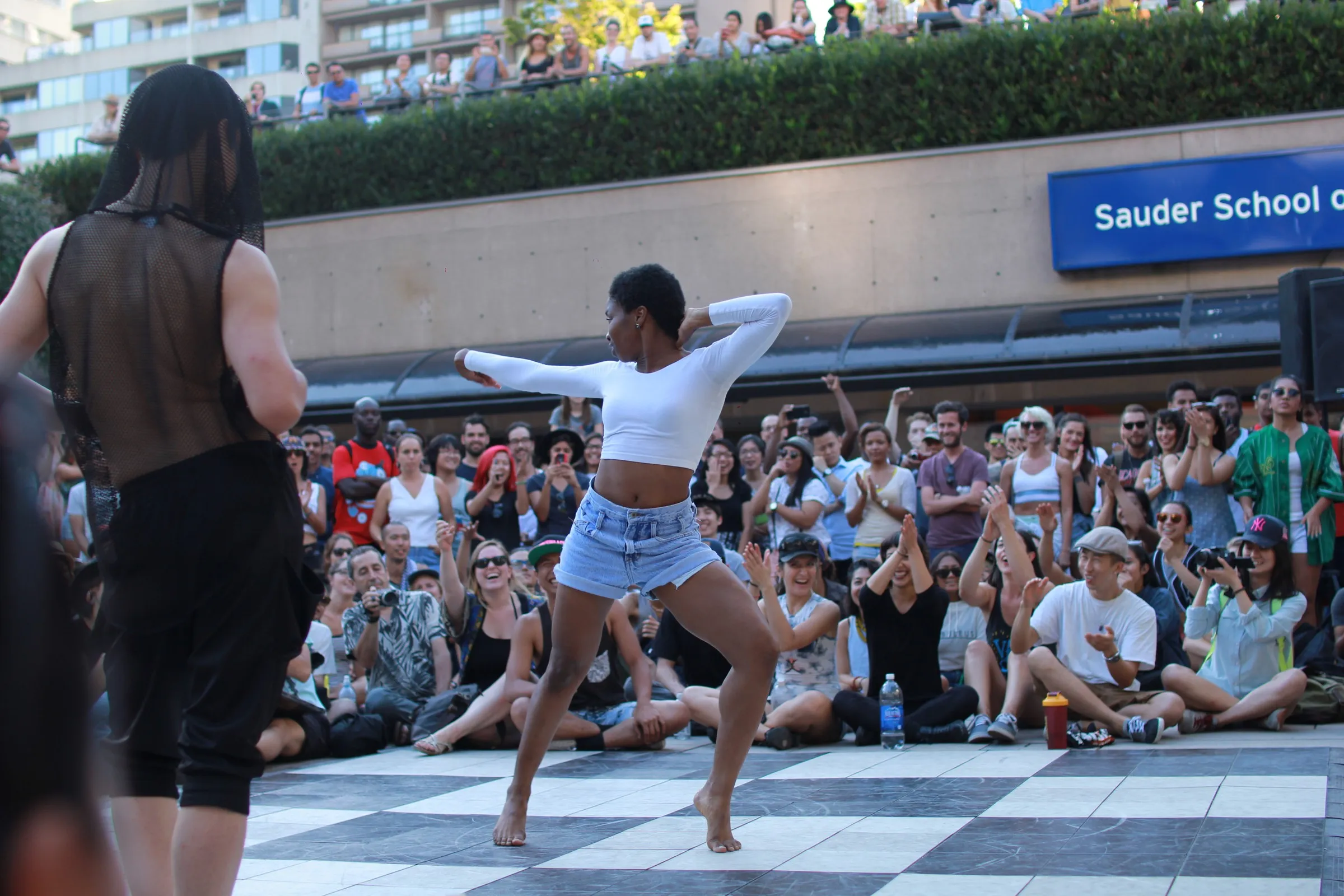 A dancer performing on a checkered floor in front of a large seated crowd outdoors