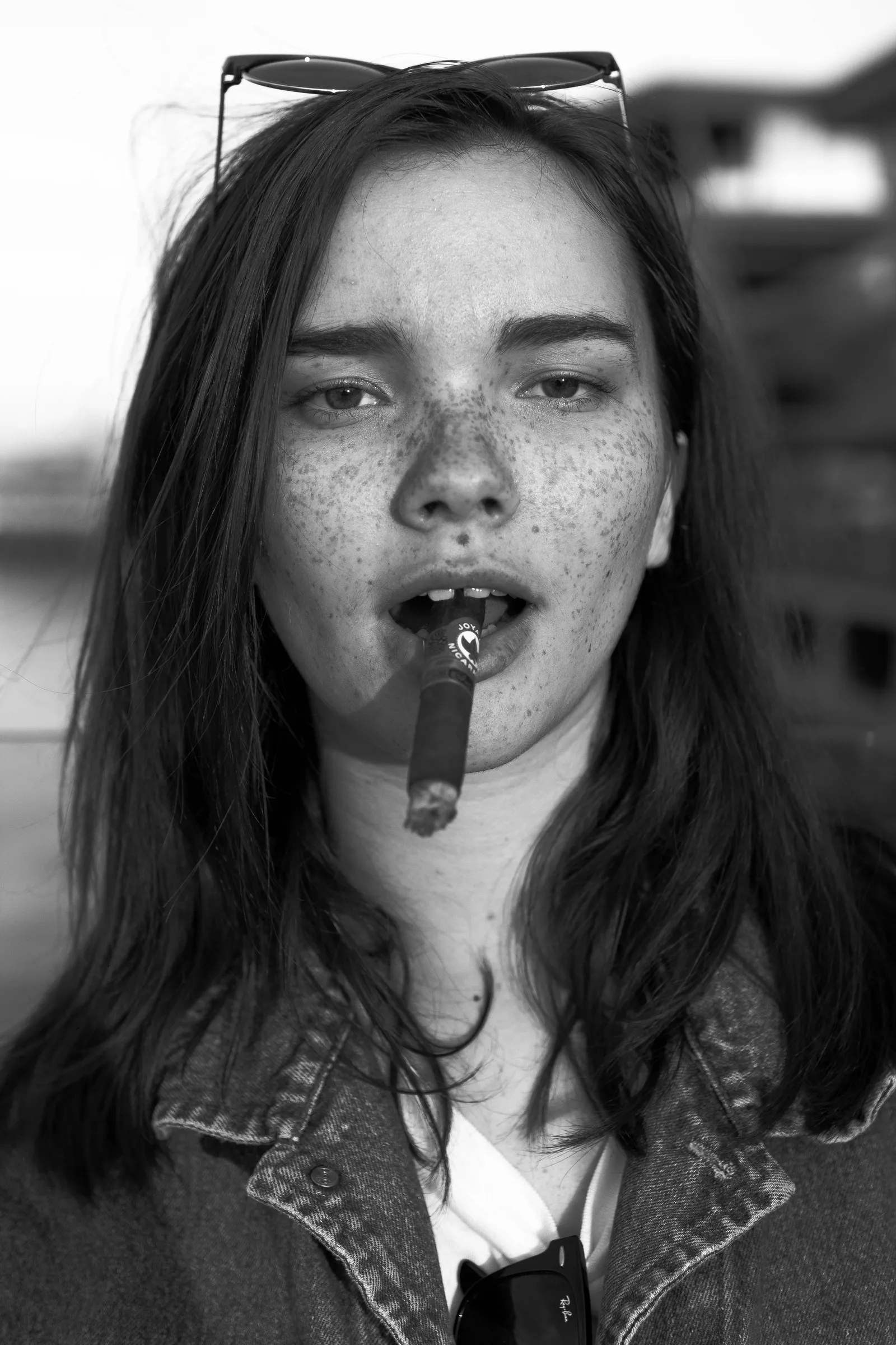 Black-and-white portrait of a freckled young woman with sunglasses on her head and a cigarette between her lips