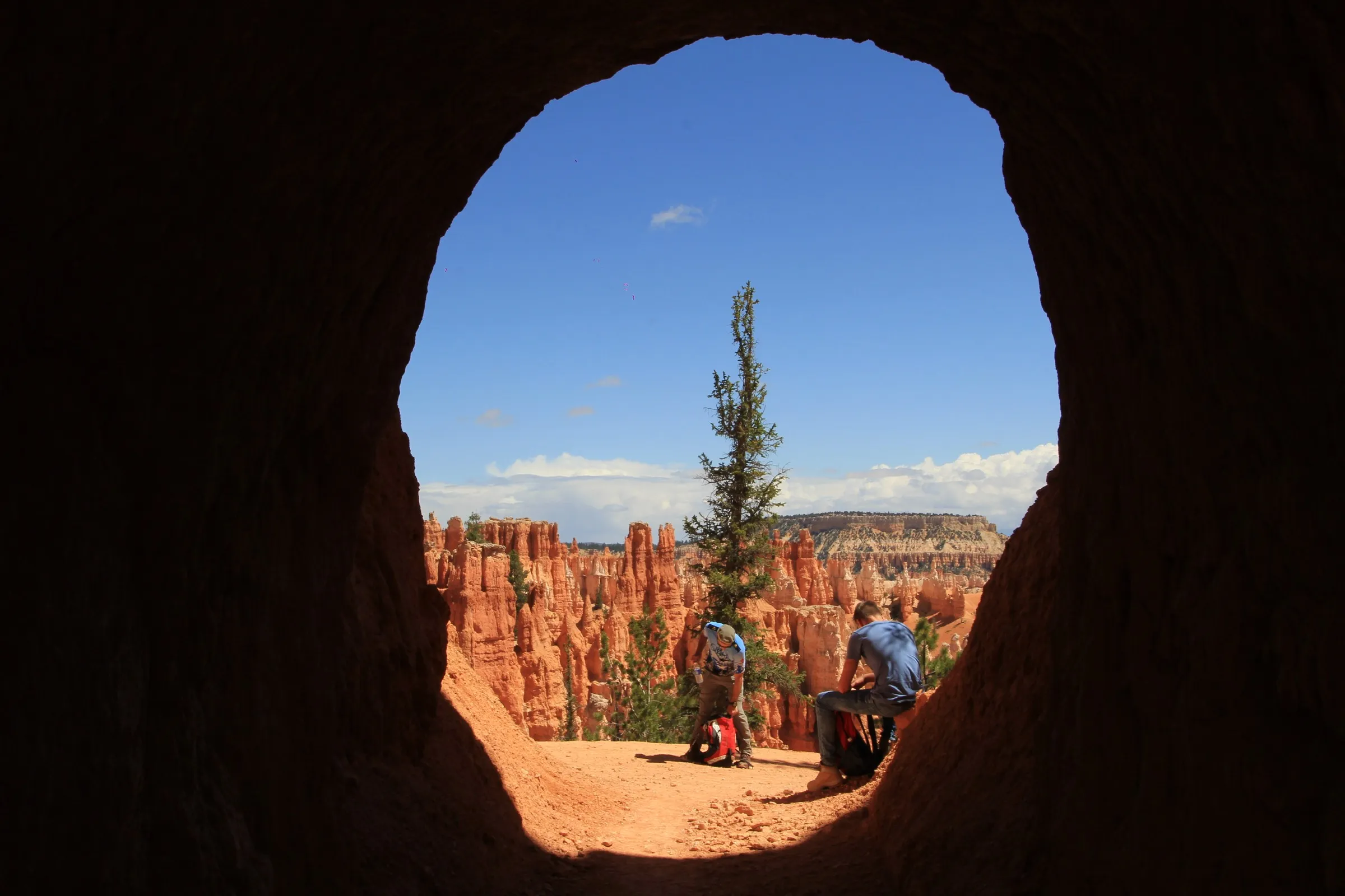 Two hikers resting in a rock arch that frames red canyon spires and a single tree