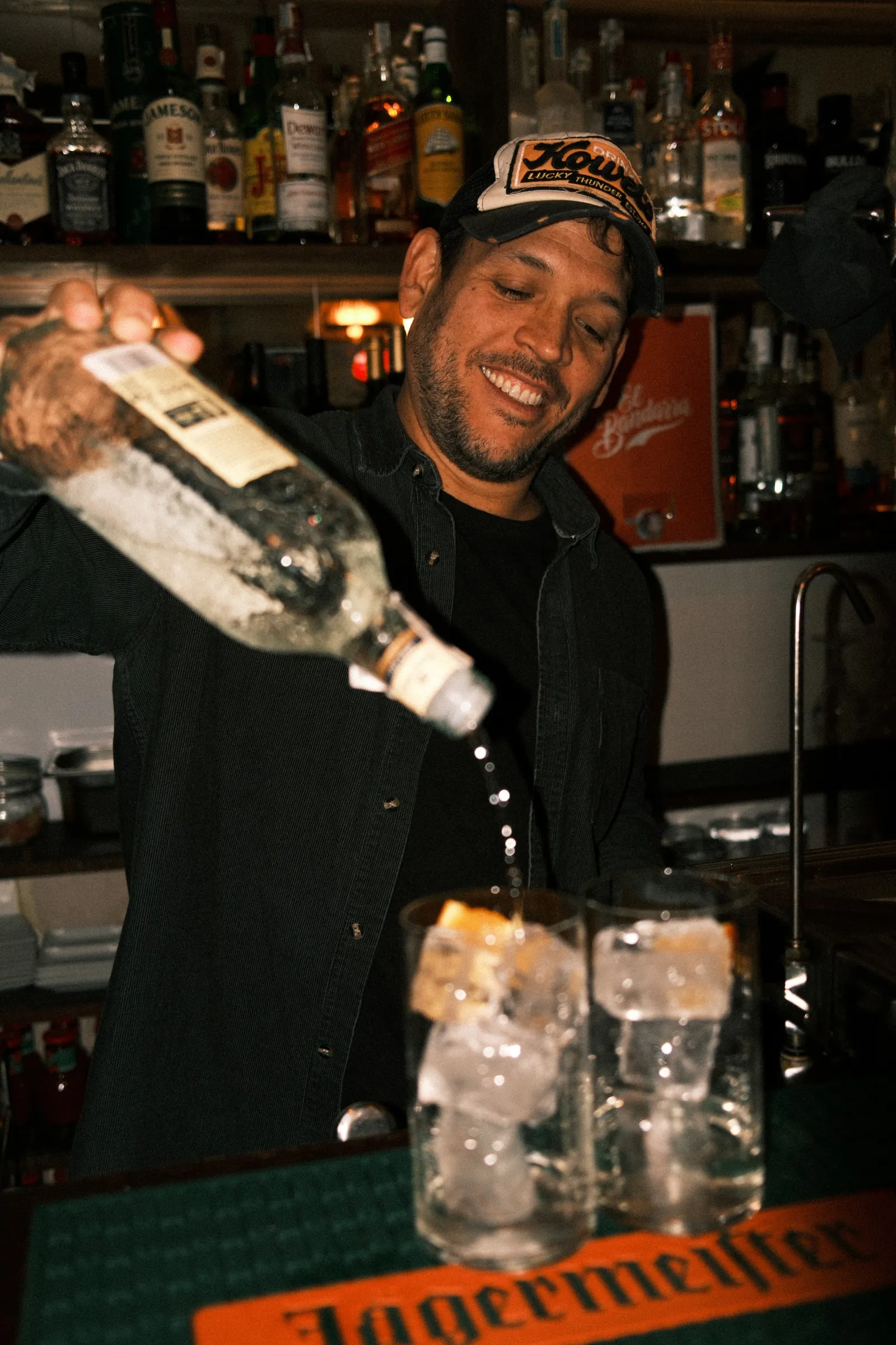 A smiling bartender pouring a drink over glasses of ice at a busy bar