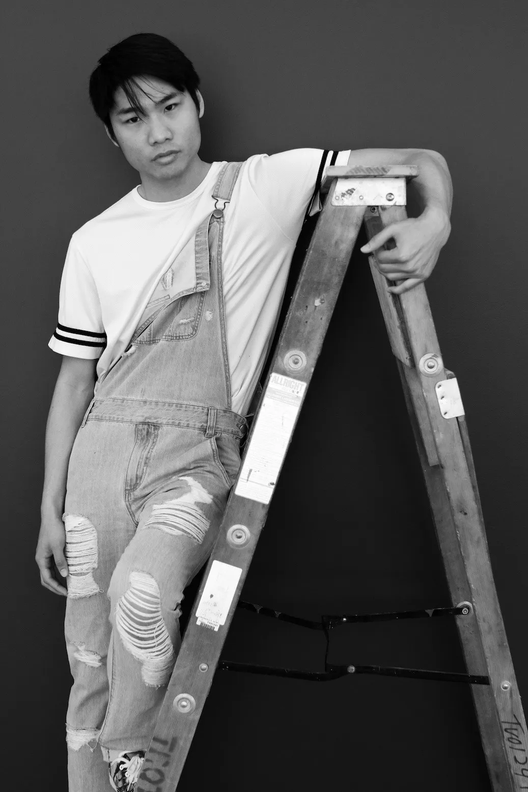 Black-and-white portrait of a young man in denim overalls leaning on a wooden ladder