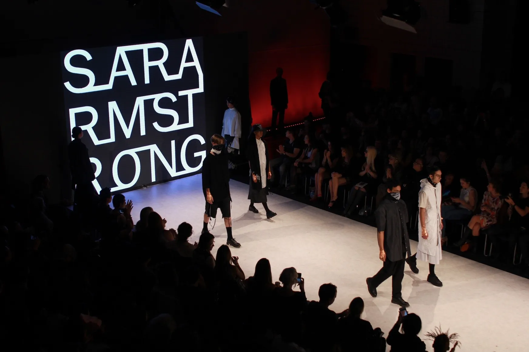 A fashion show runway seen from above, with the audience in the dark and the designer name lit on screen