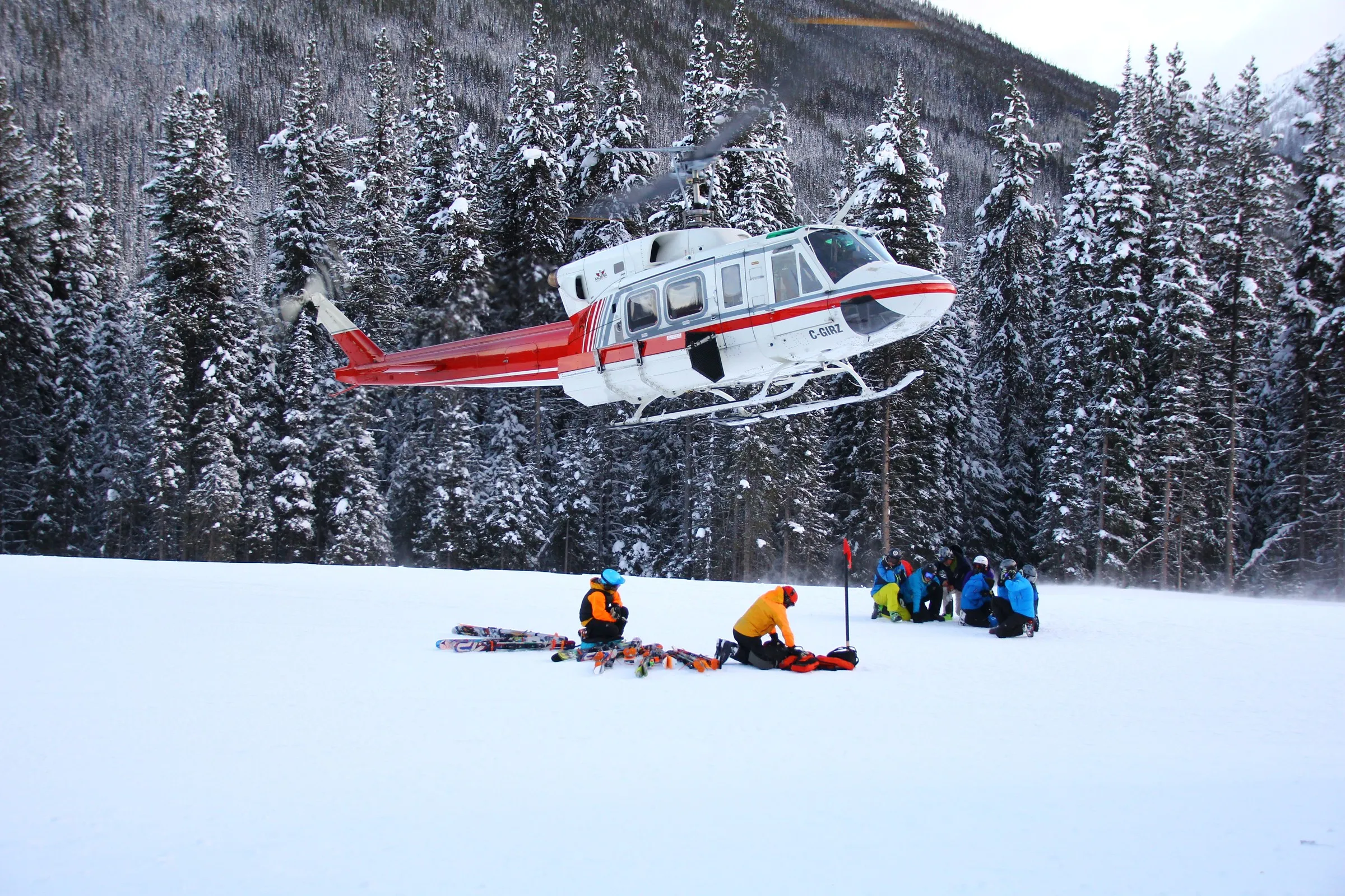 A red and white helicopter landing in a snowy forest clearing next to a group of skiers
